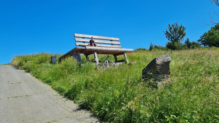 Eine große Holzbank auf einer Wiese mit einer Person darauf, umgeben von grünem Gras und einem klaren blauen Himmel.
