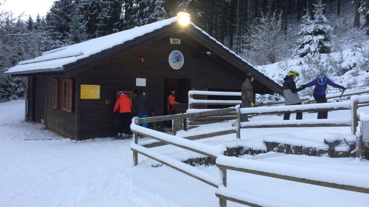 Ein Holzhäuschen im Winter, umgeben von Schnee und hohen Bäumen. Menschen stehen draußen und genießen die winterliche Landschaft.
