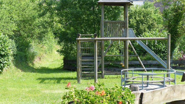 A playground with a slide and a climbing break is located in a green meadow. In the foreground, colorful flowers can be seen in a flower box.