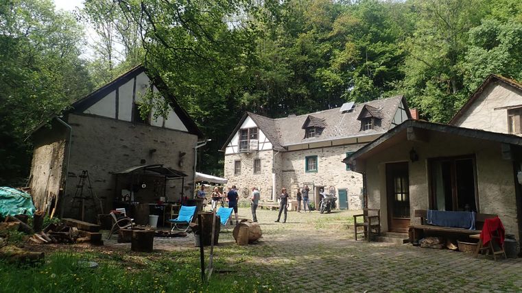 A picturesque complex with several buildings amid trees. The courtyard is equipped with seating and tools, indicating a creative atmosphere.