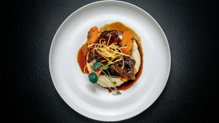 Ein kunstvoll angerichteter Teller mit zartem Fleisch, Gemüseeinlagen und einer feinen Sauce. Die Garnitur aus Kräutern und Zitronenschale verleiht dem Gericht einen frischen Touch.