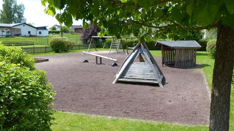 Ein Spielplatz mit Sandboden und einer Schaukel. Im Hintergrund sind grüne Wiesen und einige Gebäude zu sehen.