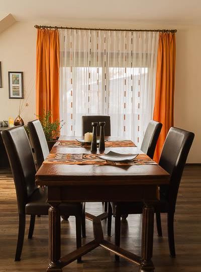Ein stilvoller Essbereich mit einem großen Holztisch und schwarzen Stühlen. Die orangefarbenen Vorhänge lassen viel Licht hereinscheinen.