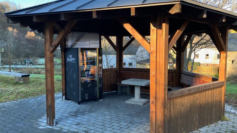 Ein überdachter Holzpavillon mit einer Sitzgelegenheit und einem Verkaufsautomaten. Die Umgebung ist grün und ruhig.