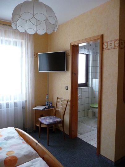 A simple bedroom with a bed, a chair, and a TV. In the background, a bathroom is visible.