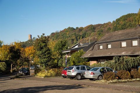Ein ruhiger Parkplatz mit mehreren Autos und Bäumen in der Umgebung. Im Hintergrund sind Hügel und ein Aussichtsturm sichtbar.