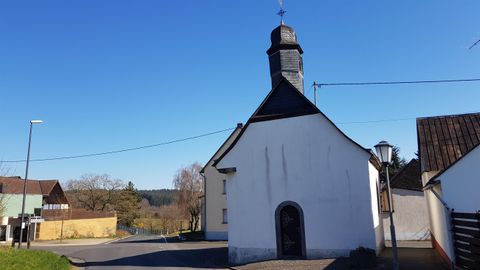 Eine kleine weiße Kirche mit einem spitzen Dach steht am Straßenrand. Der Himmel ist klar und blau, umgeben von ländlicher Szene.