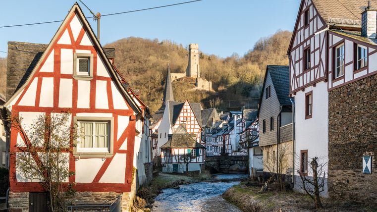 Ein malerisches Dorf mit Fachwerkhäusern am Ufer eines ruhigen Flusses. Im Hintergrund ist eine alte Burg sichtbar, die auf einem Hügel thront.