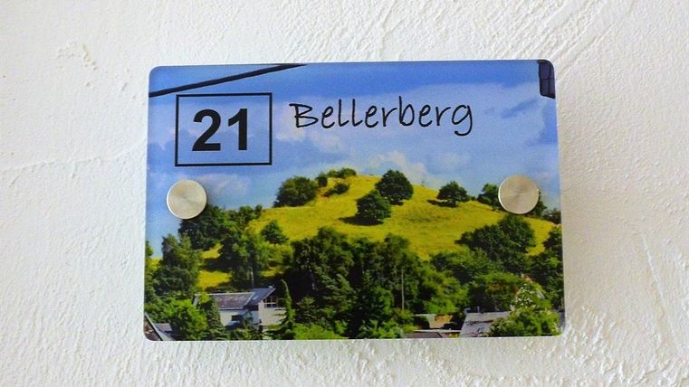 A house number with the inscription "21 Bellerberg" is displayed on a blue plaque. In the background, green hills and trees can be seen.