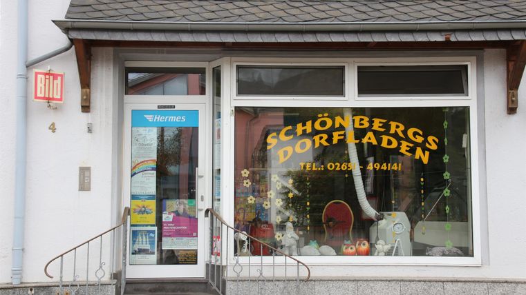 Ein kleines Geschäft mit der Aufschrift "Schönbergs Dorfladen" an der Fensterfront. Die Eingangstür ist einladend und das Geschäft bietet einen Blick auf verschiedene Waren im Schaufenster.