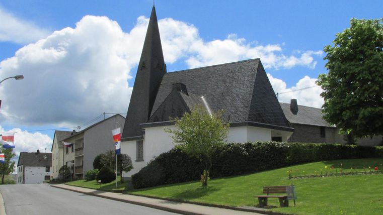 Eine Kirche mit einem spitzen Turm steht an einer ruhigen Straße. Im Hintergrund sind grüne Bäume und eine blaue Himmel mit weißen Wolken zu sehen.