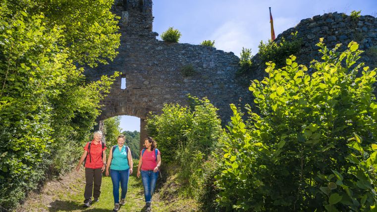 Drei Wanderer gehen durch ein Burgtor, umgeben von grünen Büschen und Bäumen, bei sonnigem Wetter.