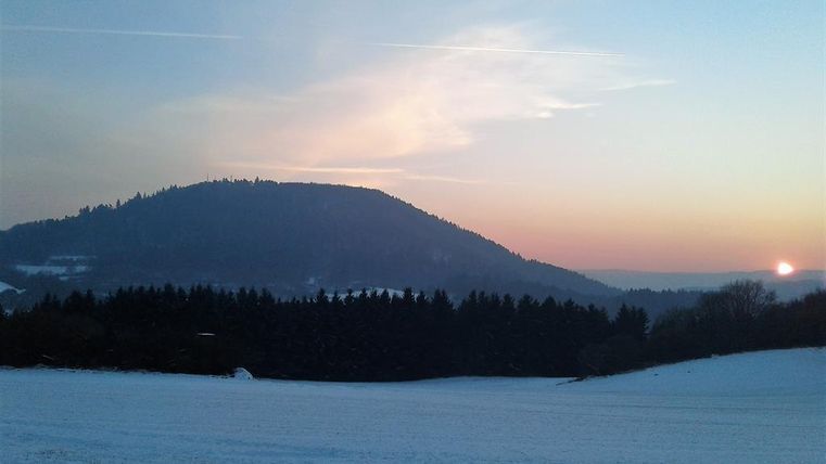 A winter landscape with a snow-covered field and a hill in the background. The sunset colors the sky in soft hues.