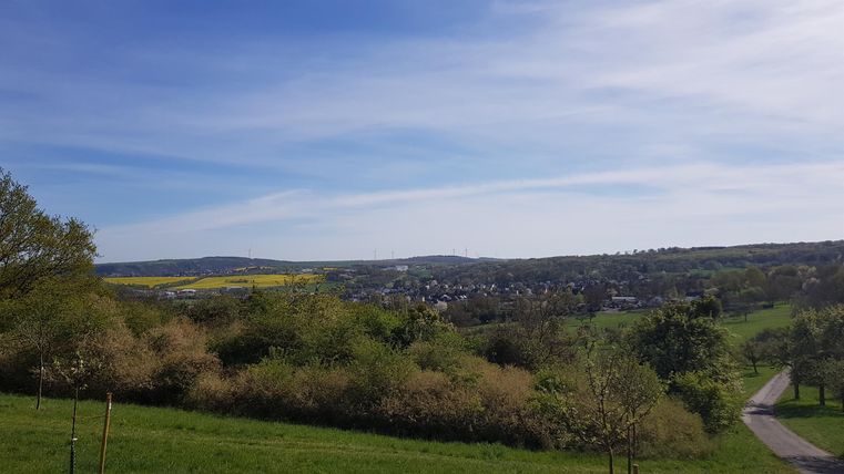 A tranquil hilly landscape with lush greenery and a clear sky. In the background, small villages are visible.