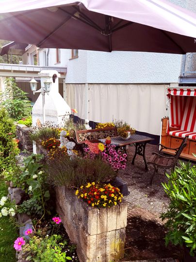 Ein gemütlicher Außenbereich mit einem Tisch und Stühlen unter einem Sonnenschirm. Bunte Blumen in verschiedenen Töpfen schmücken die Umgebung.