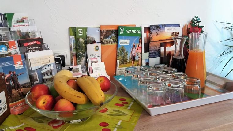 A table with a fruit bowl and information brochures for hikes. Next to it is a tray with glasses and a carafe of orange juice.