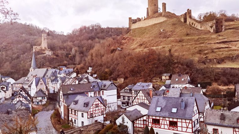 Eine malerische mittelalterliche Stadt mit Fachwerkhäusern. Im Hintergrund erhebt sich eine Burgruine auf einem Hügel.