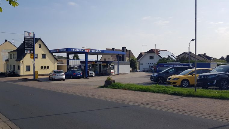 Eine Tankstelle mit mehreren Autos auf dem Parkplatz. Im Hintergrund sind einige Häuser und ein klarer Himmel zu sehen.