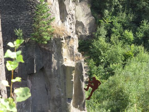 Ein Kletterer steht an einer steilen Felswand, umgeben von üppigem Grün. Die Szene vermittelt ein Gefühl von Abenteuer und Naturverbundenheit.