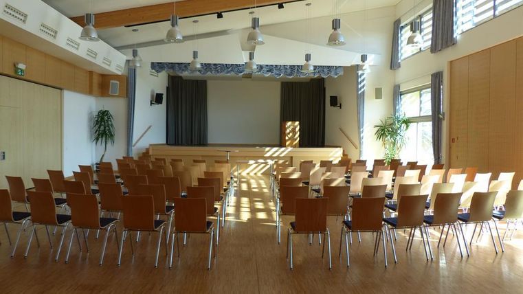 Ein gut beleuchteter Veranstaltungsraum mit Reihen von Stühlen. Im Hintergrund befindet sich eine kleine Bühne mit Vorhängen.