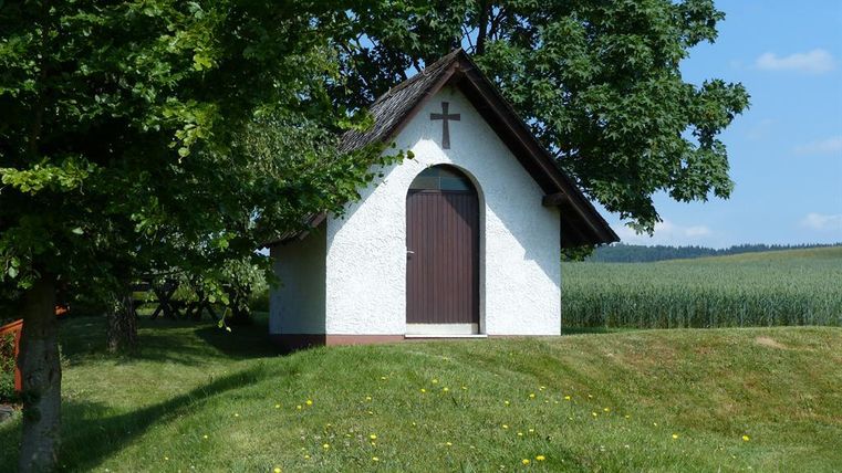 A small white chapel with a gabled roof stands on a hill. It is surrounded by green trees and meadows.