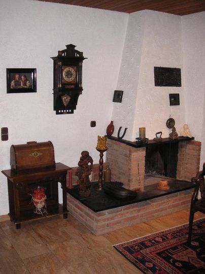 A cozy living room with a fireplace and traditional furniture. Clocks and pictures hang on the wall, creating a warm atmosphere.