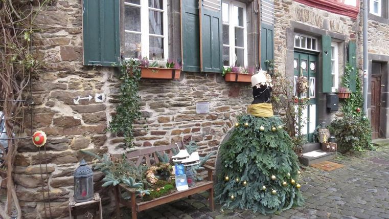 Ein historisches Haus mit grauen Steinwänden und grünen Fensterläden. Vor dem Gebäude befindet sich eine festliche Dekoration in Form eines Kleides aus Tannenzweigen.