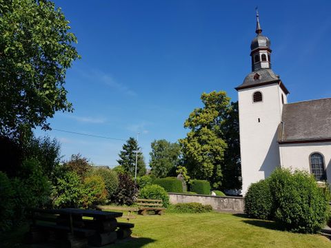 Eine malerische Kirche mit einem hohen Turm steht inmitten eines schönen Gartens. Grüne Bäume und Bänke laden zum Verweilen ein.