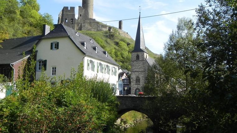 A picturesque landscape with a castle on a hill and historic buildings in the foreground. A stream flows under a stone bridge.