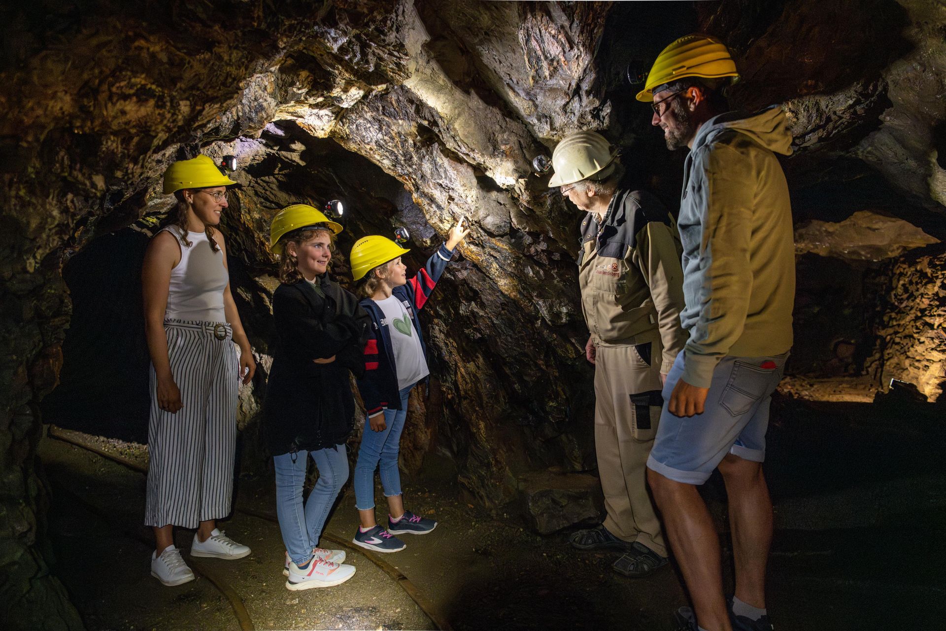 Eine Gruppe von Menschen mit Helmen erkundet einen Dunkelraum in einer Mine. Eine Person zeigt auf etwas in der Wand, während andere aufmerksam zuschauen.
