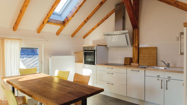 Eine moderne Küche mit Holzdeckenbalken und großen Fenstern. Der Essbereich hat einen Holztisch mit gelben Stühlen.