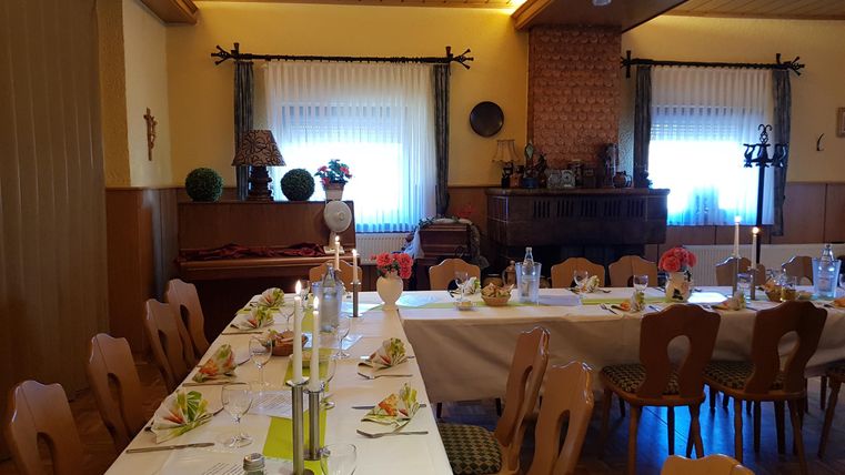 Ein schön dekorierter Essraum mit einem langen Tisch und Stühlen. Die Tische sind für ein Festmahl vorbereitet, mit Gläsern und Besteck.