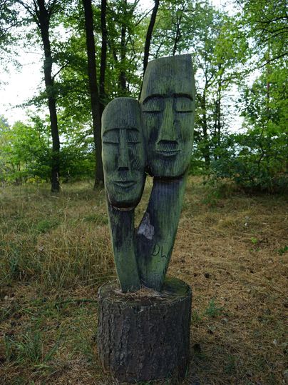 Eine Holzskulptur mit zwei stilisierten Gesichtern steht in einem Wald. Der Boden ist mit Gras bedeckt und umgeben von Bäumen.