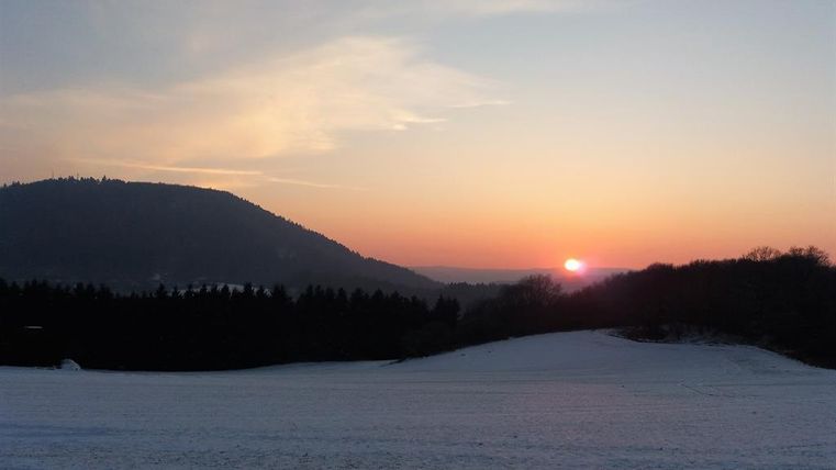 A picturesque winter landscape with a snow-covered field and a beautiful sunset behind the mountains. The sky displays soft colors of orange and blue.