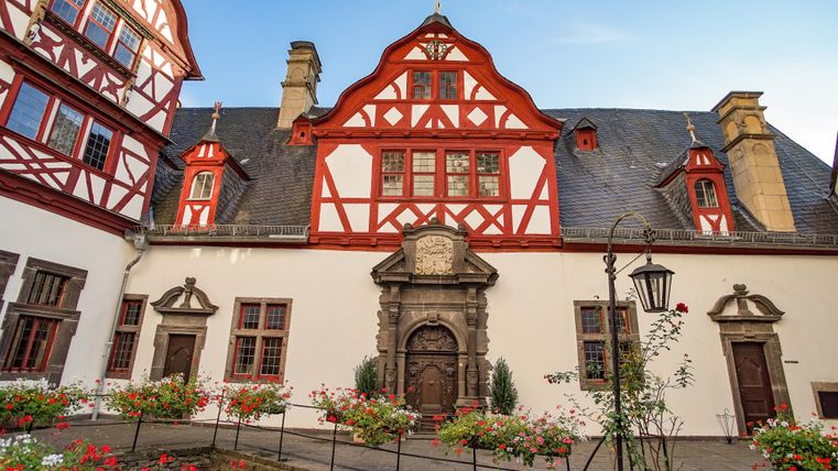 A historic building with a distinctive red half-timbering. The courtyard is surrounded by colorful flowers.