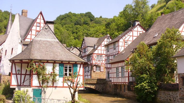 Ein malerisches Dorf mit Fachwerkhäusern und einer kleinen Brücke über einen ruhigen Fluss. Die grünen Hügel im Hintergrund tragen zur idyllischen Atmosphäre bei.