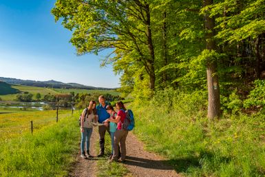 Familie mit Wanderkarte in der Hand auf einem Feldweg mit Aussicht und Waldbereich