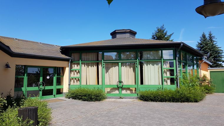 Ein helles Gebäude mit grünen Fenstern und einem schlichten Dach. Umgeben von Büschen und einem klaren blauen Himmel.