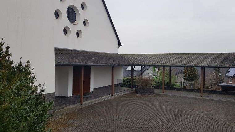 A modern building with a white exterior plaster and large round windows. In the foreground, there is a covered area and a paved floor.