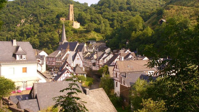 Eine malerische Dorfansicht mit Fachwerkhäusern und einer Kirche. Im Hintergrund ragt eine alte Burg über den bewaldeten Hügel.