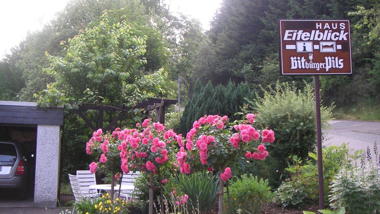 An inviting house with colorful flowers and a sign that says "Eifelblick House." The surroundings are green and well-kept.