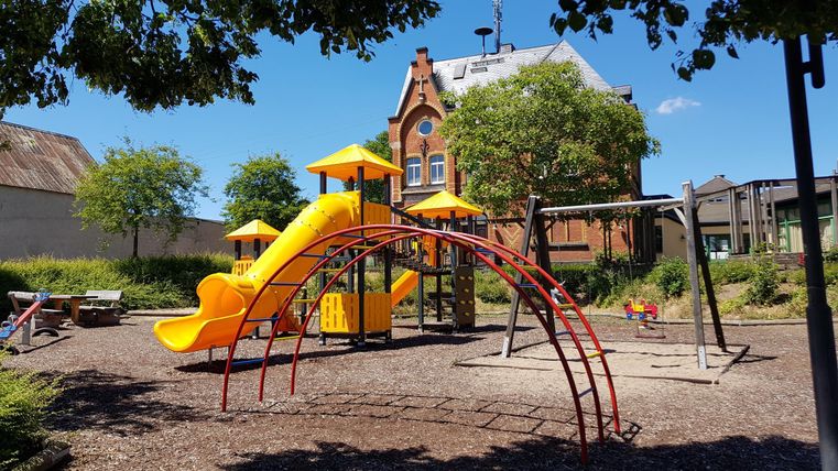 Ein bunter Spielplatz mit einer Rutsche und Klettergerüsten. Im Hintergrund steht ein charmantes Gebäude und es ist sonnig.