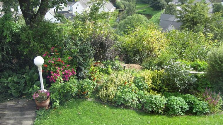 Ein schöner Garten mit bunten Blumen und einer gepflegten Rasenfläche. Im Hintergrund sind kleine Häuser und Bäume in einer grünen Landschaft zu sehen.