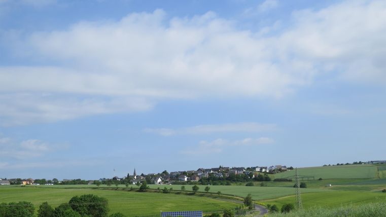 Eine malerische Landschaft mit sanften Hügeln und einem kleinen Dorf im Hintergrund. Der Himmel ist blau mit einigen Wolken.