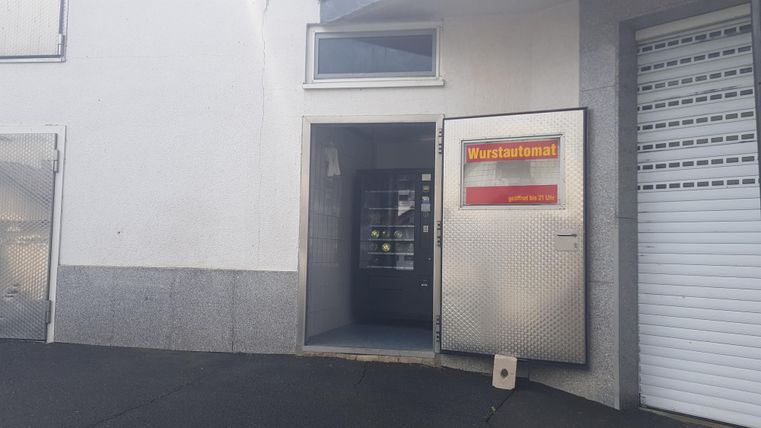 Ein Eingang zu einem Wurststand in einem grauen Gebäude. Die Tür ist offen und das Schild mit der Aufschrift "Wurststand" ist deutlich sichtbar.