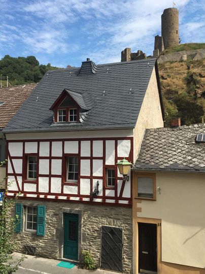 Ein charmantes Fachwerkhaus mit roten Akzenten und einem grünen Eingang. Im Hintergrund sieht man eine alte Burgruine auf einem Hügel.