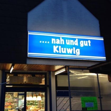 Ein Geschäft mit einem leuchtenden Schild, das "nah und gut Kluwig" zeigt. Die Eingangstür ist sichtbar und die Umgebung wirkt einladend.