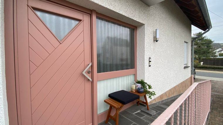 Eine Eingangstür mit einem modernen, rosa Design. Vor der Tür steht eine kleine Sitzgelegenheit mit Pflanzen.
