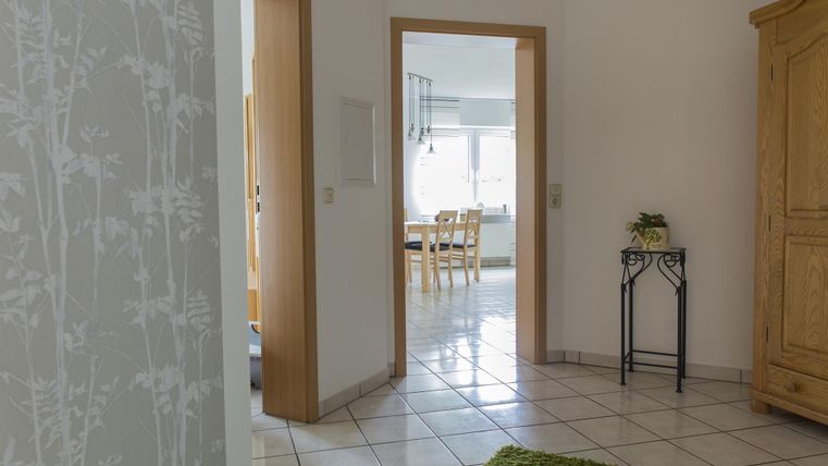 A bright hallway with tiled flooring and a green carpet mat. Through the door, a dining area with a wooden table can be seen.