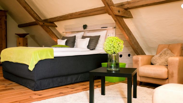 A cozy attic room with a large bed and bright decorations. Next to the bed is a black table with a vase of flowers and a beige armchair.